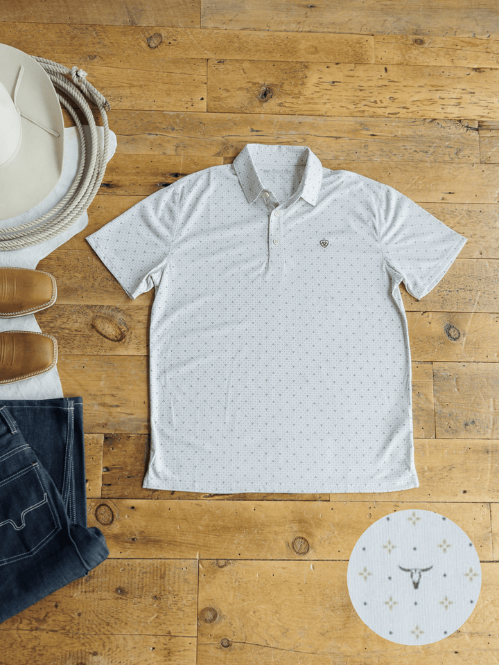 The Dupree Ariat Men's Southwest Polo from Savannah Sevens western life{&}style, shown in white with a subtle rodeo-themed pattern, lies on a wooden floor beside cowboy gear. An inset spotlights its moisture-wicking feature and detailed icons.