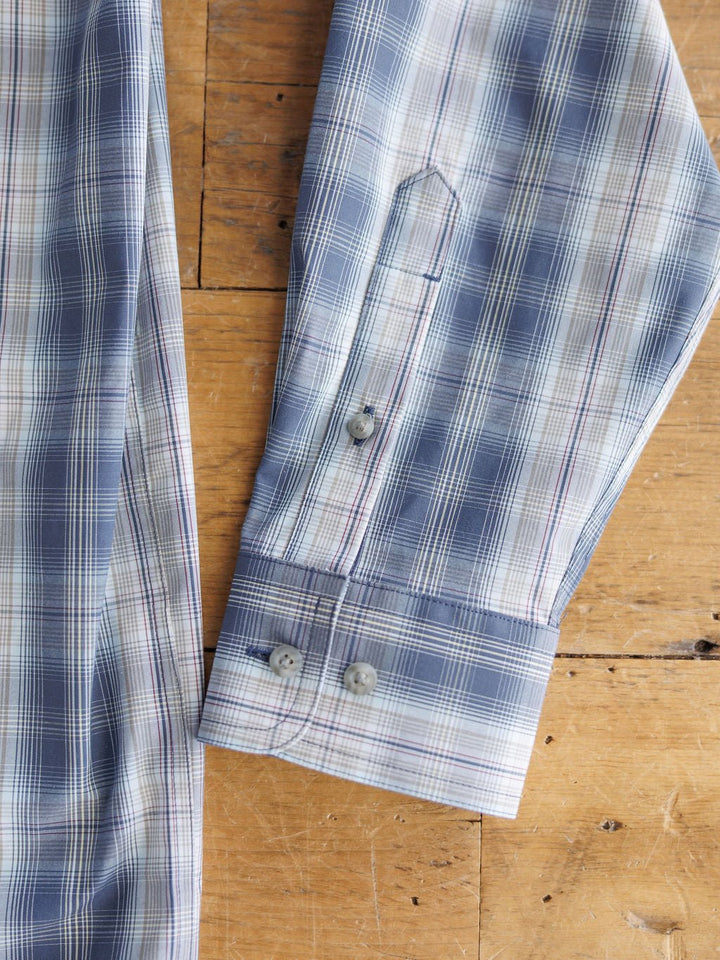 A close-up of the Durand Ariat Men's Plaid Button Up by Savannah Sevens western life{&}style shows its blue and white check cuff with two buttons on a wooden surface. Moisture-wicking fabric ensures all-day comfort.