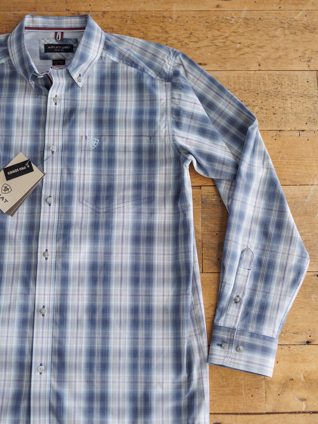The Durand Ariat Men's Plaid Button Up from Savannah Sevens western life{&}style, in blue and white with long sleeves, lies flat on wood. It features sun protection, moisture wicking, button-up design, and tags by the collar.