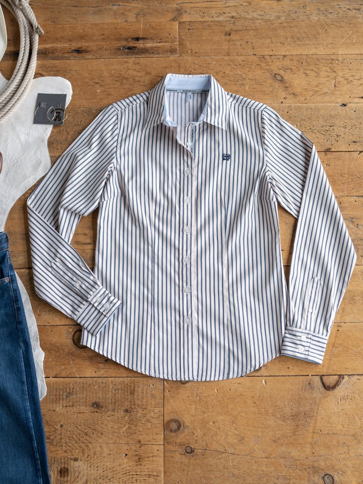 The Durant (S-XXL) by Savannah Sevens western life{&}style—a white women’s fitted shirt with blue vertical stripes—is neatly displayed on a wooden floor, with glimpses of a white tote bag and blue jeans at the edges.