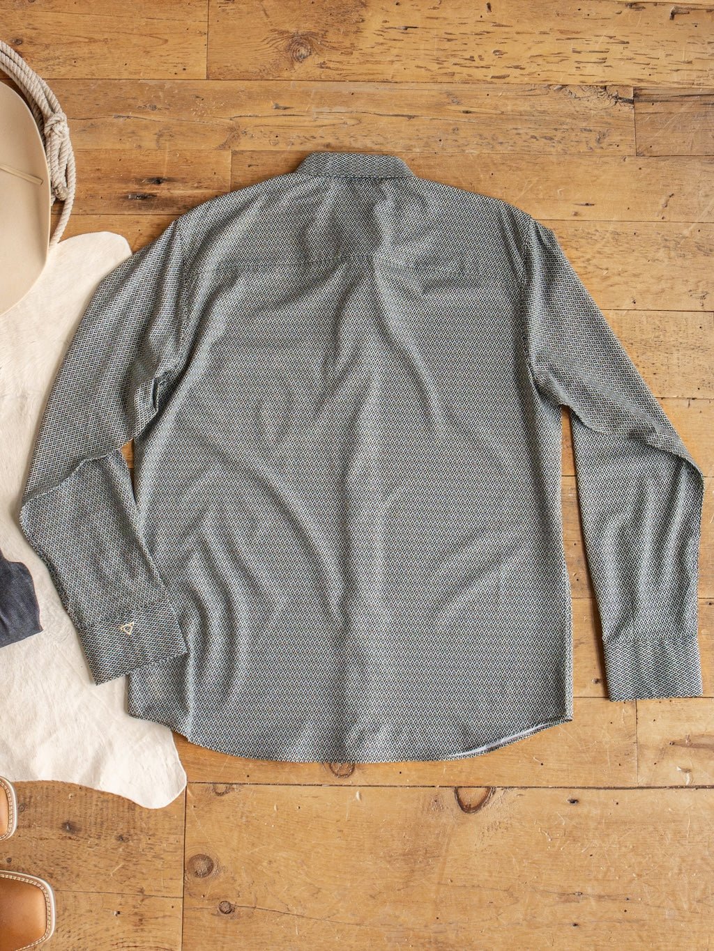 The Durham Wrangler Men's Southwest Button Up from Savannah Sevens western life{&}style is laid flat, back facing up, on a wooden floor beside a white rug and partially visible tan shoes. This men’s long sleeve shirt showcases a southwest pattern and features moisture-wicking fabric for added comfort.