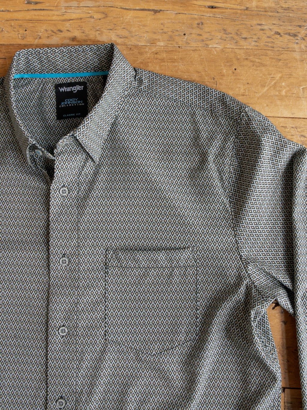 The Durham Wrangler Men's Southwest Button Up by Savannah Sevens western life{&}style, featuring a small black and white geometric diamond design inspired by classic southwest patterns and a front pocket, is laid out on a wooden surface.