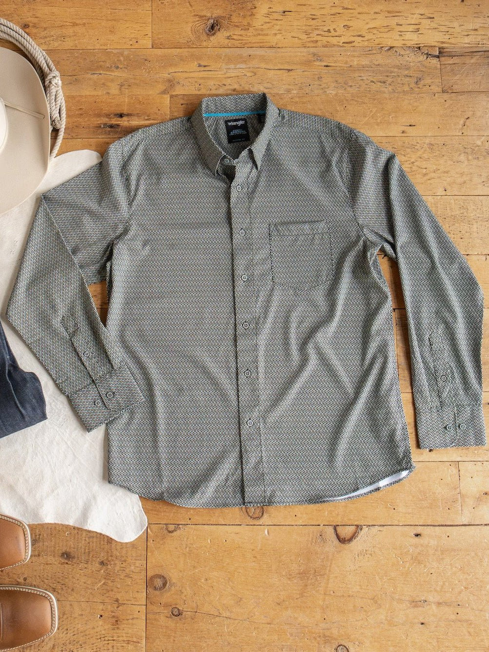 The Durham Wrangler Men's Southwest Button Up by Savannah Sevens western life{&}style, featuring a green southwest pattern and chest pocket, is displayed flat on a wooden floor beside a white hat and brown boots.