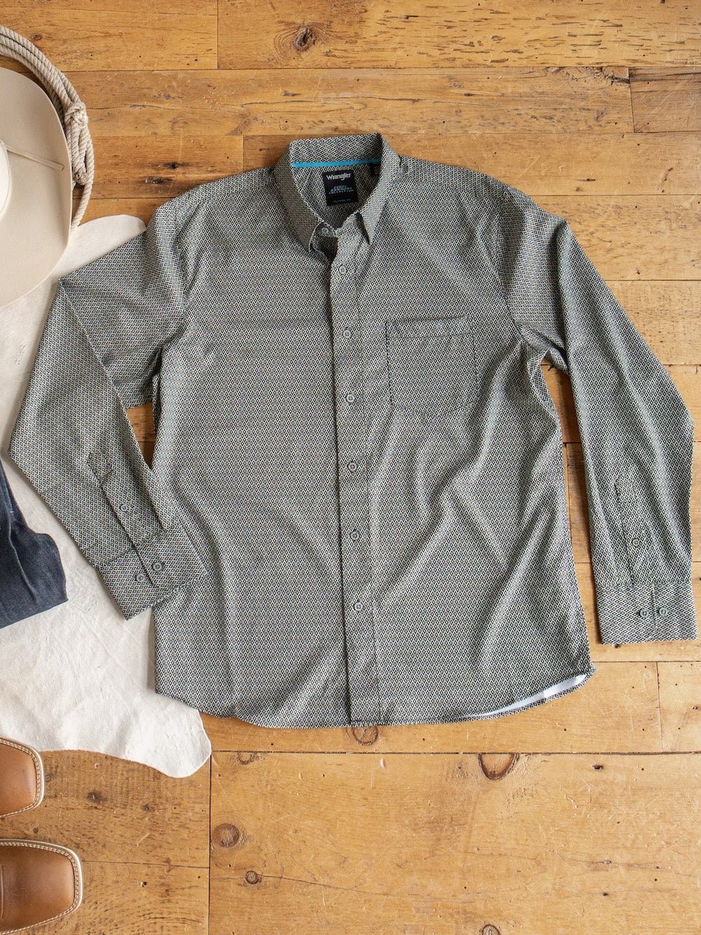 The Durham Wrangler Men's Southwest Button Up by Savannah Sevens western life{&}style, featuring a green southwest pattern and chest pocket, is displayed flat on a wooden floor beside a white hat and brown boots.