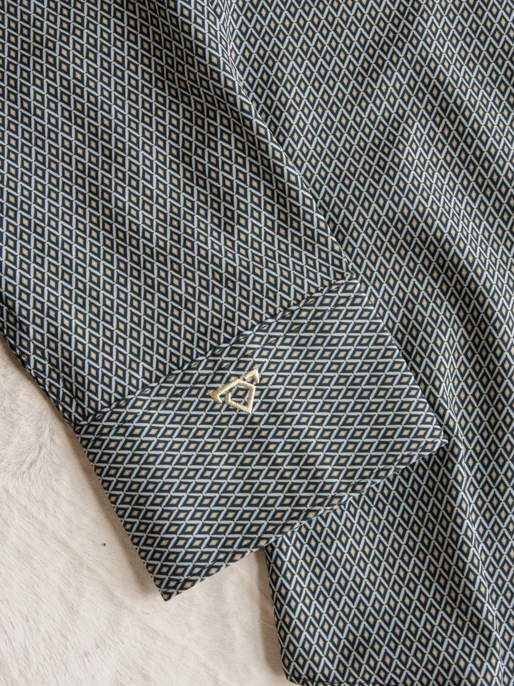 A close-up of the Durham Wrangler Men's Southwest Button Up cuff from Savannah Sevens western life{&}style, showcasing its black and white geometric diamond pattern and accented with a gold cufflink, lying on a light, textured surface.