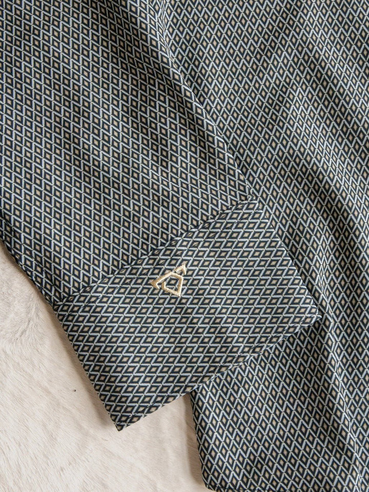 A close-up of the Durham Wrangler Men's Southwest Button Up cuff from Savannah Sevens western life{&}style, showcasing its black and white geometric diamond pattern and accented with a gold cufflink, lying on a light, textured surface.
