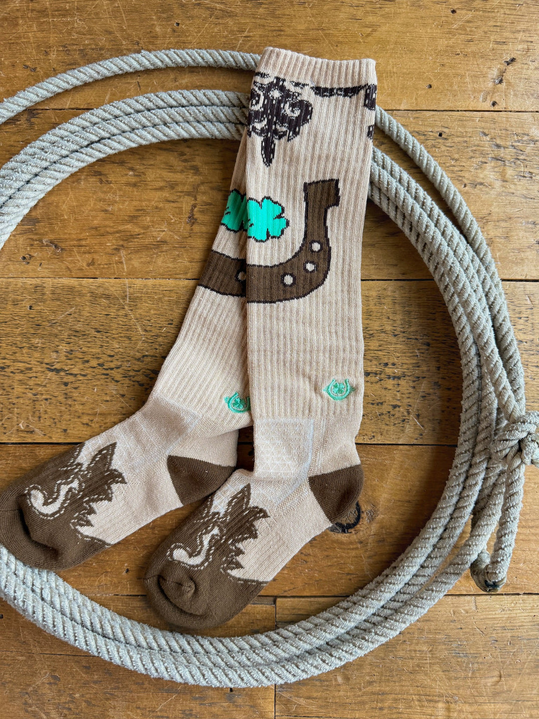 A pair of Dust Kicker Crew Socks by Savannah Sevens western life{&}style, featuring a tan color with Viking ship and dragon designs, lie inside a coiled rope on a wooden floor. These breathable socks are crafted to ensure comfort throughout the day.