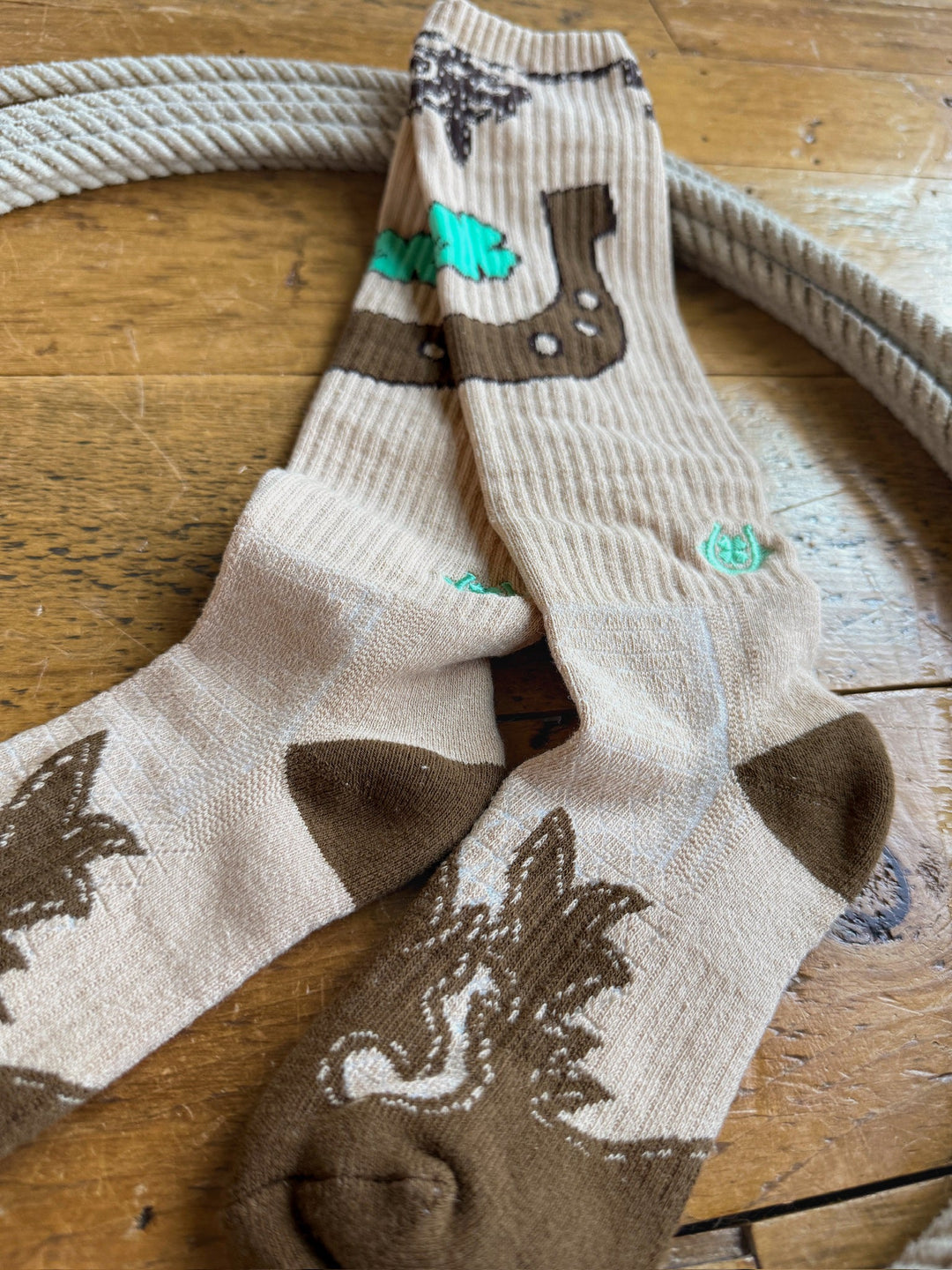 A pair of Savannah Sevens western life{&}style Dust Kicker Crew Socks in tan, featuring brown and green bear and leaf designs, lies crossed on a wooden floor next to a coiled rope. These breathable socks are crafted with odor-fighting hemp for all-day comfort.