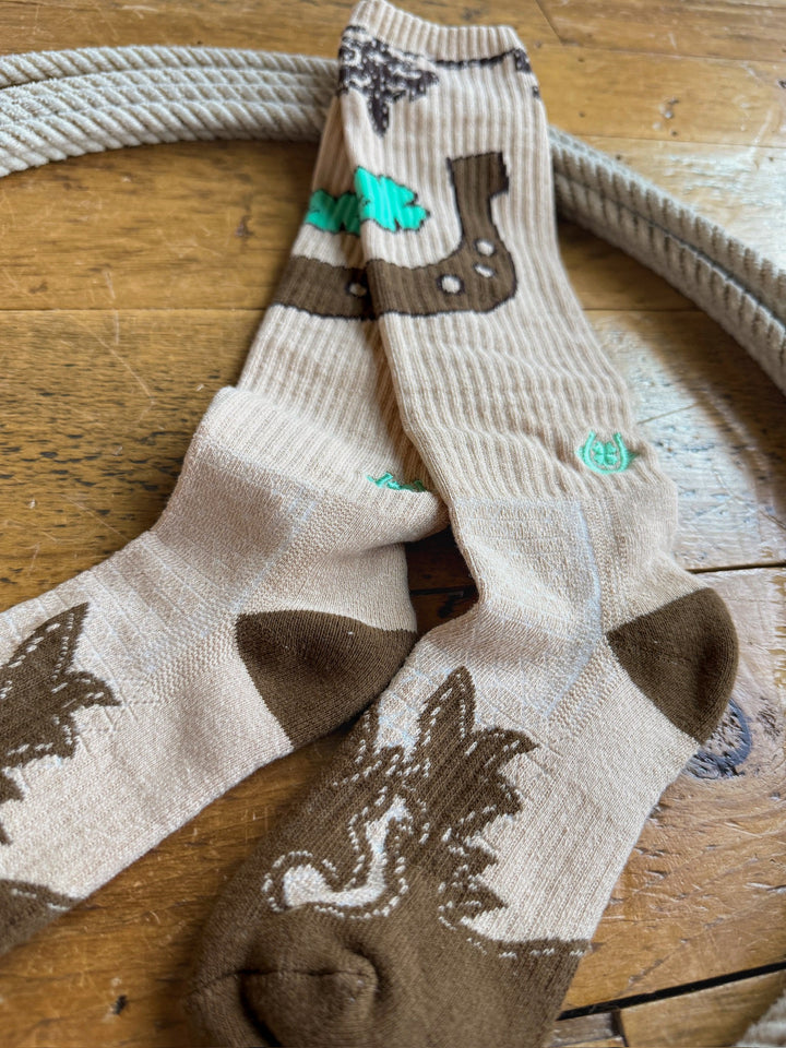 A pair of Savannah Sevens western life{&}style Dust Kicker Crew Socks in tan, featuring brown and green bear and leaf designs, lies crossed on a wooden floor next to a coiled rope. These breathable socks are crafted with odor-fighting hemp for all-day comfort.