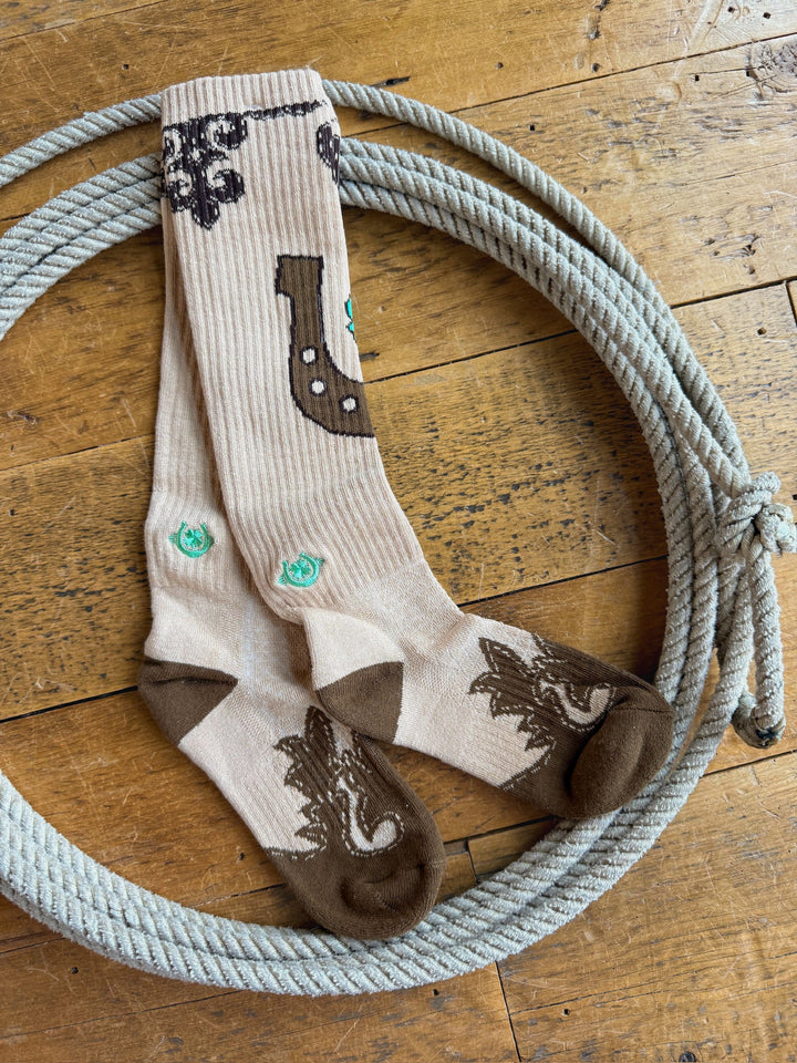 Dust Kicker Crew Socks by Savannah Sevens western life{&}style, featuring beige and brown horse-themed designs, are displayed atop a coiled rope on a wooden floor. Made with breathable fabric and odor-fighting hemp, these performance crew socks provide lasting comfort.