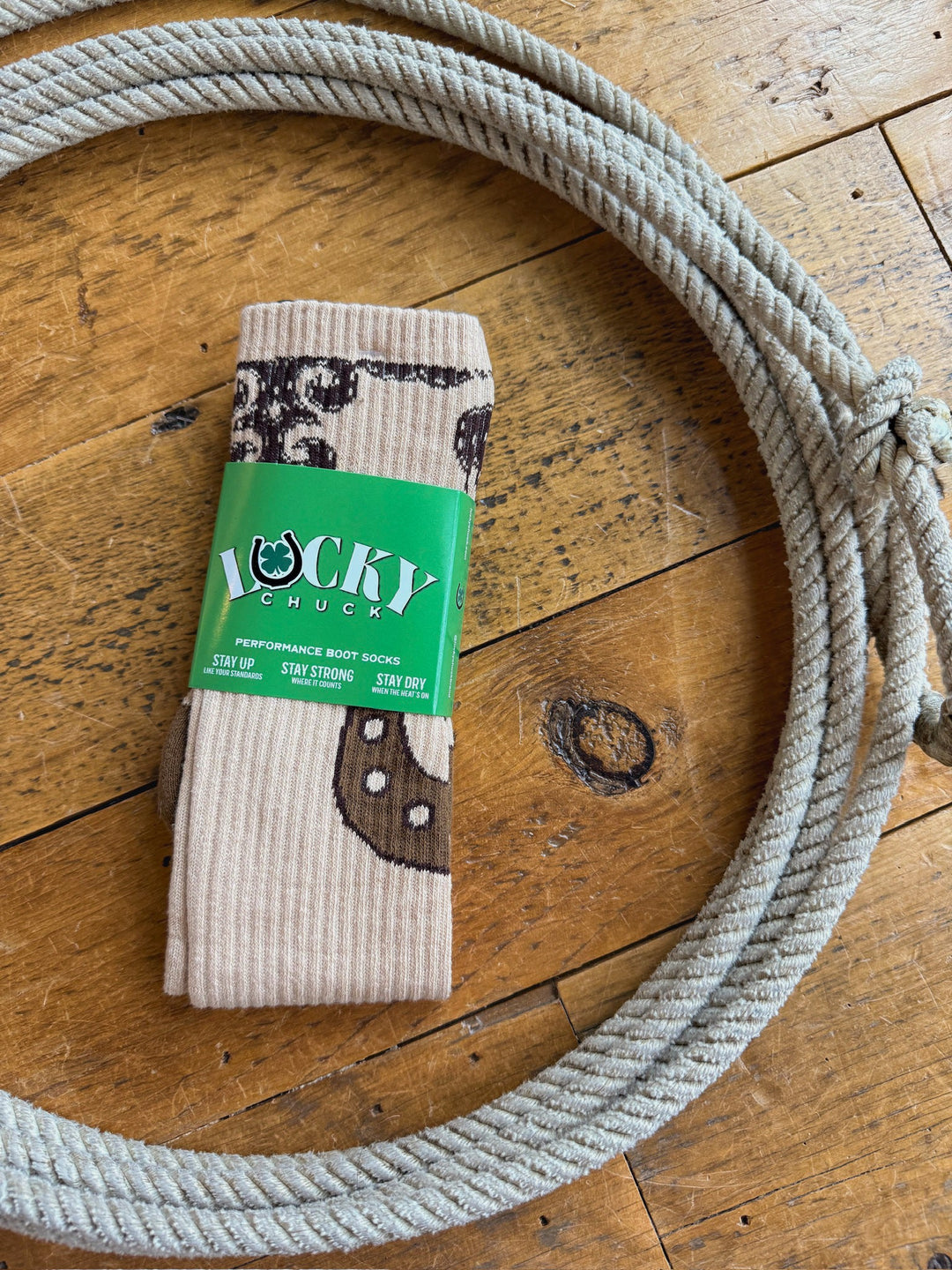 A pair of tan Dust Kicker Crew Socks by Savannah Sevens western life{&}style, featuring a green label, is placed on a wooden floor and surrounded by a coiled rope. Designed for comfort and performance, these breathable socks are perfect for western lifestyles.
