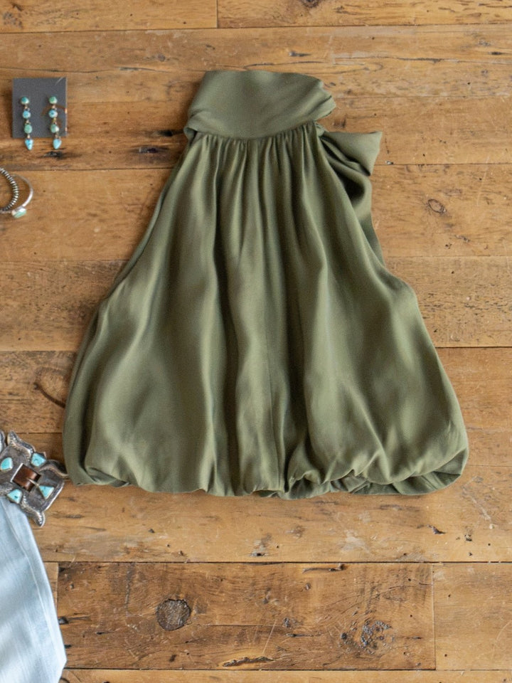The Duval Olive Bubble Hem Sleeveless Top by Savannah Sevens western life{&}style lies flat on a wooden surface, its gathered and billowy design highlighted. Silver jewelry and part of a gray garment are partially visible at the edges, while an adjustable neckline tie enhances the versatility of this olive high neck top.