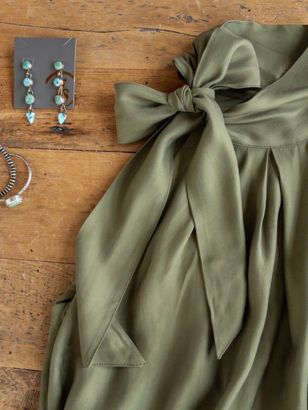 The Duval Olive Bubble Hem Sleeveless Top by Savannah Sevens western life{&}style is styled with turquoise drop earrings and a matching bracelet, all displayed on a wooden surface.