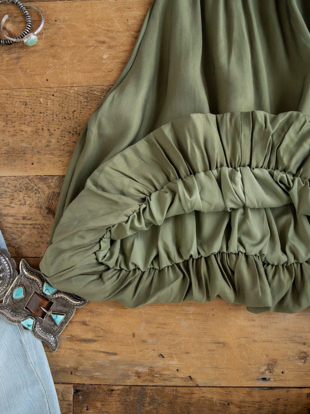 A close-up of the Duval Olive Bubble Hem Sleeveless Top by Savannah Sevens western life{&}style on a wooden surface, with part of an adjustable turquoise-and-silver belt and light blue denim fabric visible in the corner.