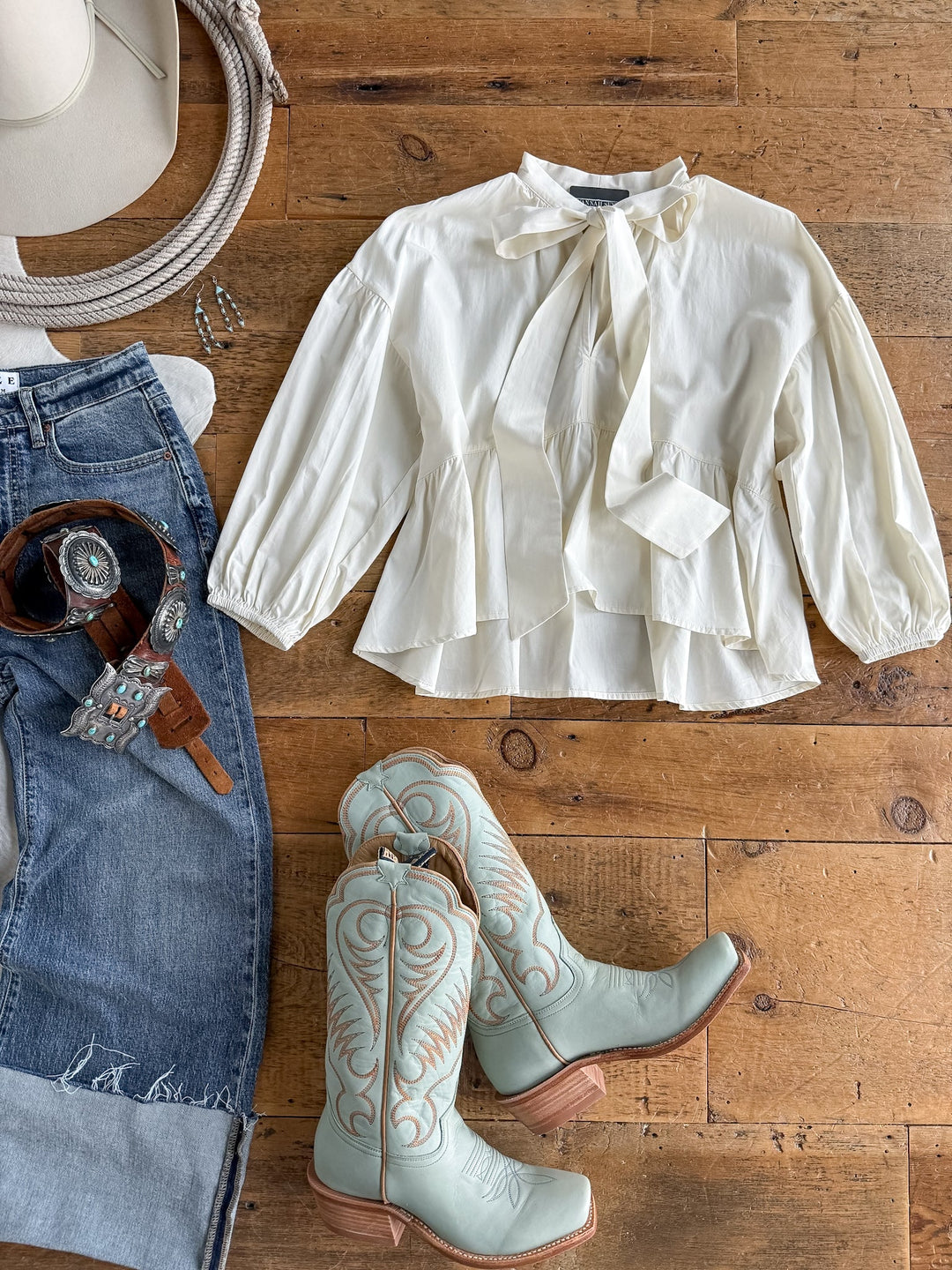 Flat lay of the Eastbrook Cream Cotton Blouse by Savannah Sevens western life{&}style, paired with light blue cowboy boots, blue jeans, a western belt featuring a large buckle, and a white cowboy hat on a wooden floor.