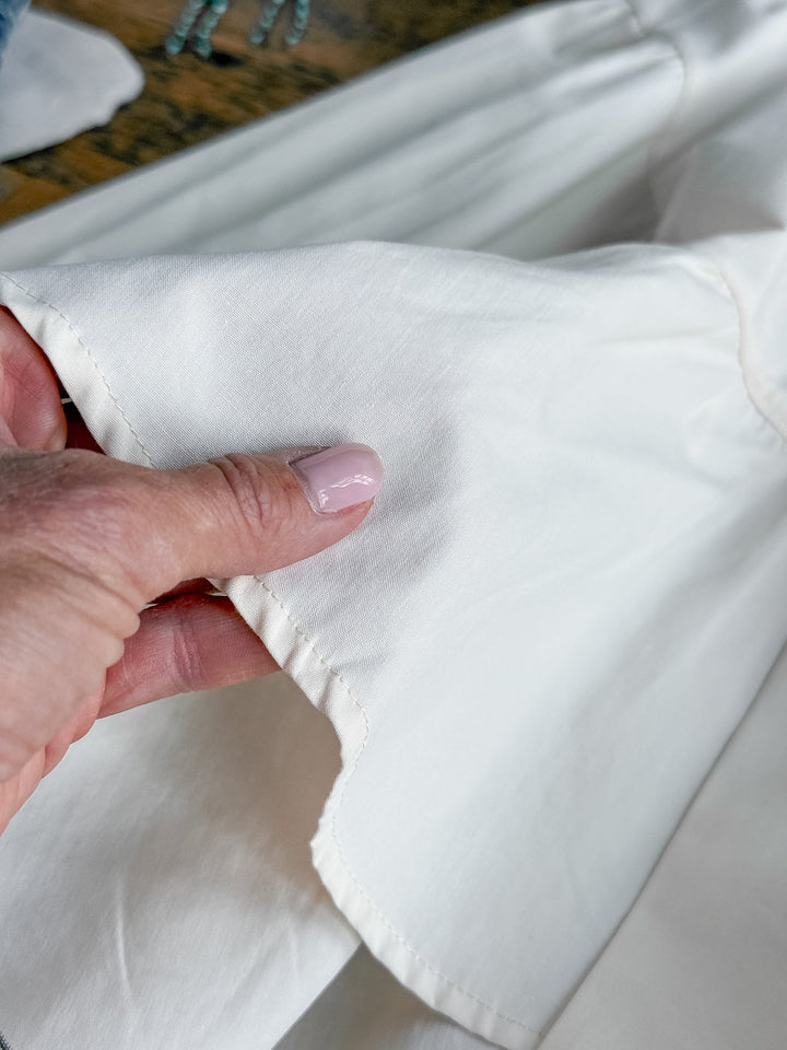 A hand with light pink nail polish holds the edge of the Eastbrook Cream Cotton Blouse by Savannah Sevens western life{&}style, highlighting its texture and fine stitching detail.