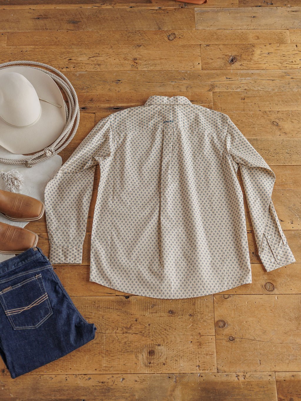The Easton Ariat Men's Southwest Button Up by Savannah Sevens western life{&}style, a light-colored shirt with a small pattern, is laid face-down on a wooden floor with a white cowboy hat, brown boots, and folded blue jeans nearby.