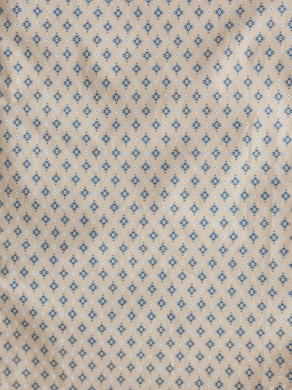 Close-up of beige fabric from the Easton Ariat Men's Southwest Button Up by Savannah Sevens western life{&}style, featuring a small blue and black geometric floral grid pattern with a slightly wrinkled, textured look.