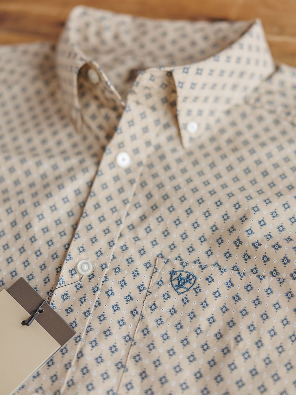 Easton Ariat Men's Southwest Button Up by Savannah Sevens western life{&}style features a beige color with a small blue geometric pattern, white buttons, and a blue embroidered chest logo, shown on a wooden surface.