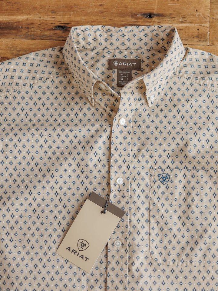 The Easton Ariat Men's Southwest Button Up by Savannah Sevens western life{&}style, in a cream color with a blue diamond pattern, is displayed on wood with a branded Ariat tag attached to the top buttonhole.