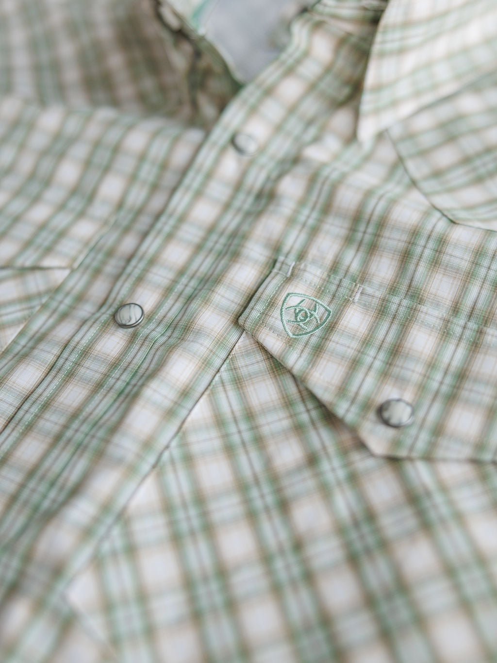 Close-up of the Eastwood Ariat Men's Plaid Button Up by Savannah Sevens western life{&}style in green and beige, featuring snap buttons and a small embroidered logo on the pocket flap.