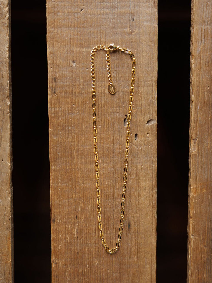 The Eckley Necklace by Savannah Sevens western life{&}style is a gold choker featuring small rectangular paperclip links and an adjustable clasp, displayed vertically on a wooden surface with visible grain and gaps.
