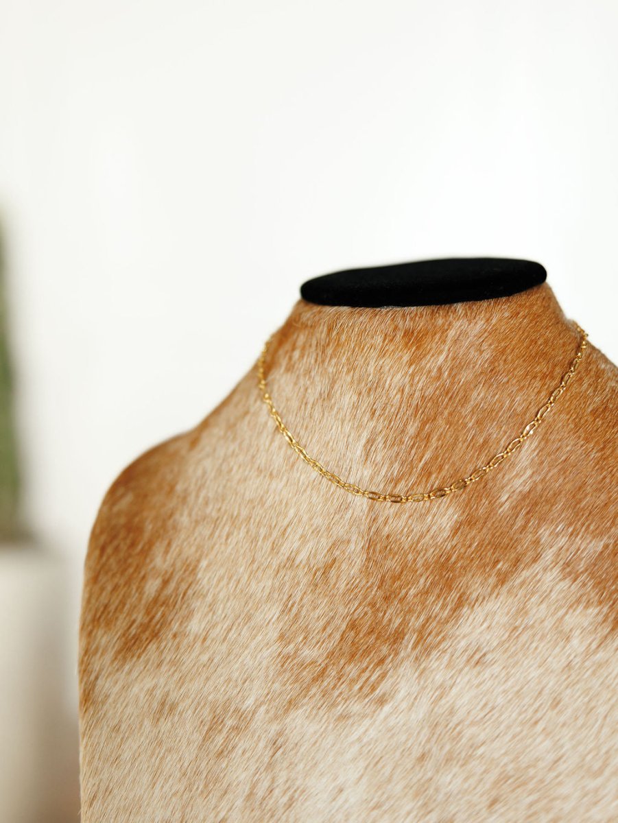 The Eckley Necklace by Savannah Sevens western life{&}style features a delicate 18K gold-filled paperclip design, displayed on a tan and white cowhide bust with a softly blurred light background.