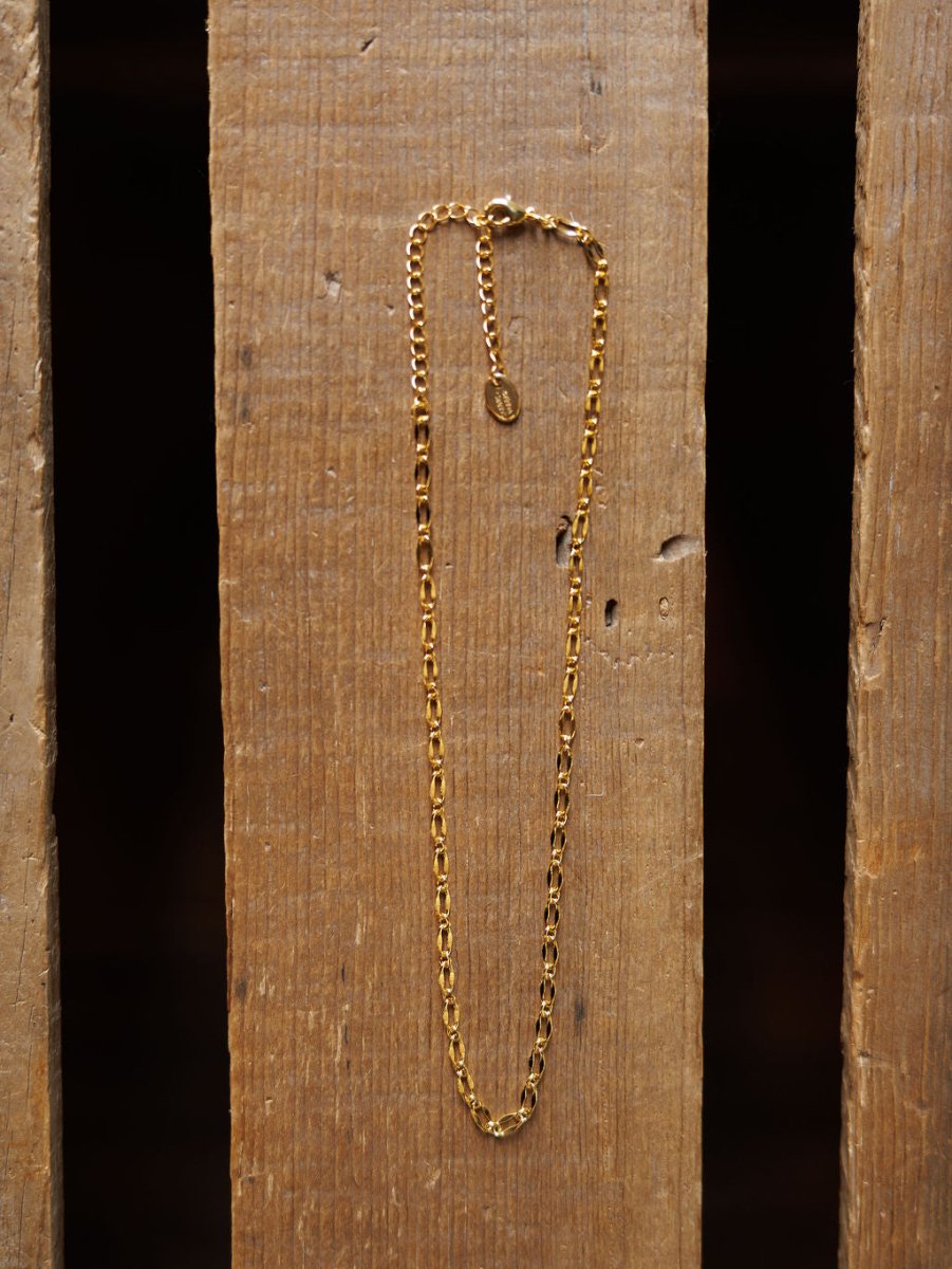 The Eckley Necklace by Savannah Sevens western life{&}style is a gold choker featuring small rectangular paperclip links and an adjustable clasp, displayed vertically on a wooden surface with visible grain and gaps.