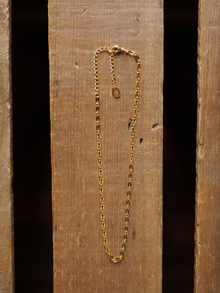 The Eckley Necklace by Savannah Sevens western life{&}style is a gold choker featuring small rectangular paperclip links and an adjustable clasp, displayed vertically on a wooden surface with visible grain and gaps.