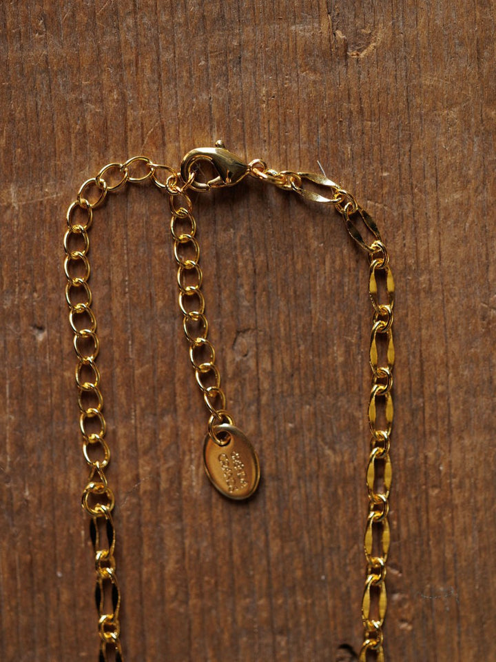 A close-up of the Eckley Necklace by Savannah Sevens western life{&}style, featuring an 18K gold filled paperclip design, lobster clasp, extender chain, and small oval tag on a textured wooden surface.