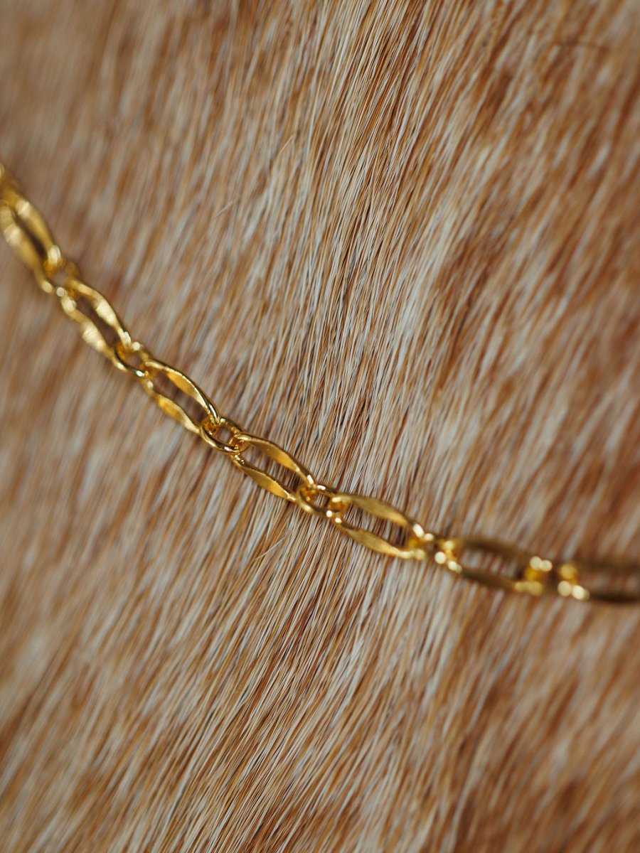 The Eckley Necklace by Savannah Sevens western life{&}style features an 18K gold filled chain resting on light brown cowhide with white streaks, highlighting a striking contrast between the gleaming metal and the soft, textured background.