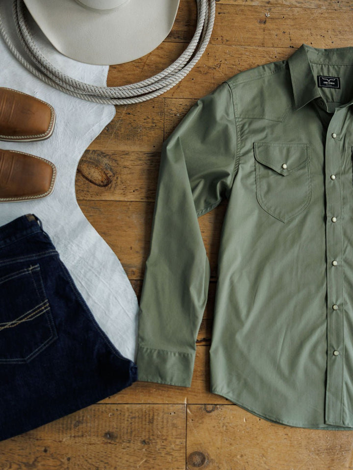 A neatly arranged flat lay featuring The Edgerton {Men's} sage green shirt by Savannah Sevens western life{&}style, dark jeans, tan cowboy boots, a white cowboy hat, and a coiled rope on a wooden floor.