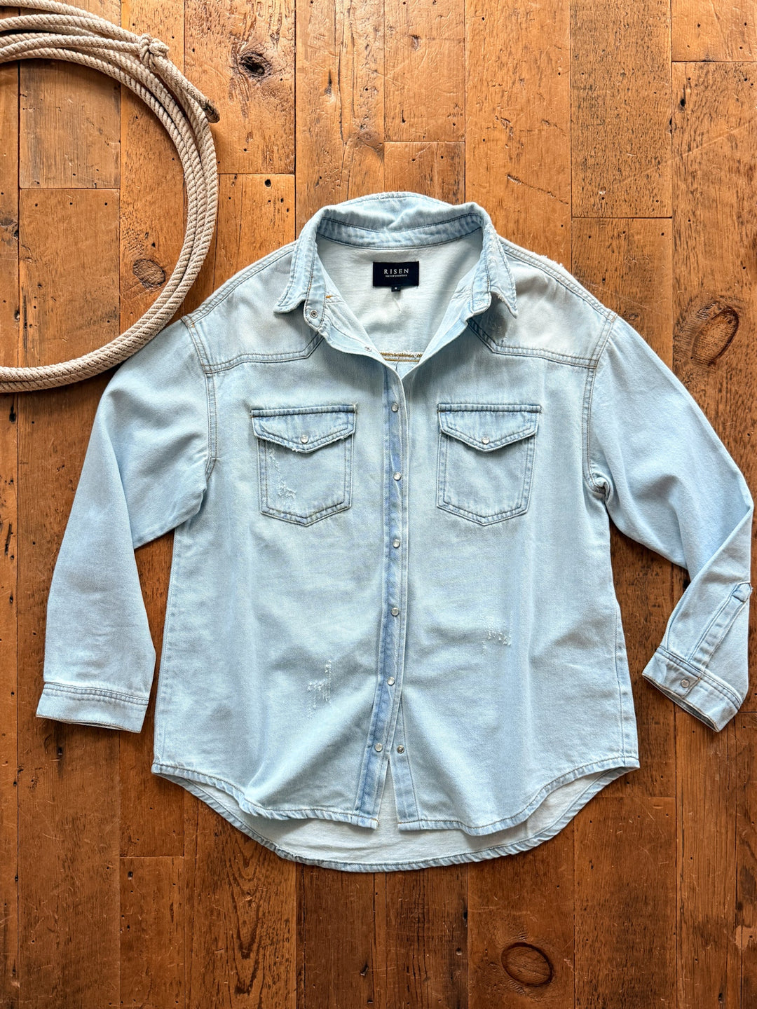 Savannah Sevens western life&style Eldorado Denim Pearl Snap oversized light-wash denim button-down shirt with distressed pearl snaps, two chest pockets, and light distressing on a wooden surface next to a coiled rope.