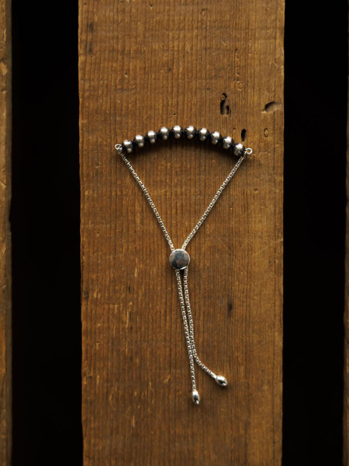 The Elida Bracelet by Savannah Sevens western life{&}style, featuring round dark Navajo pearls and an adjustable sterling silver chain with sliding clasp, is displayed vertically on a rustic wooden surface.