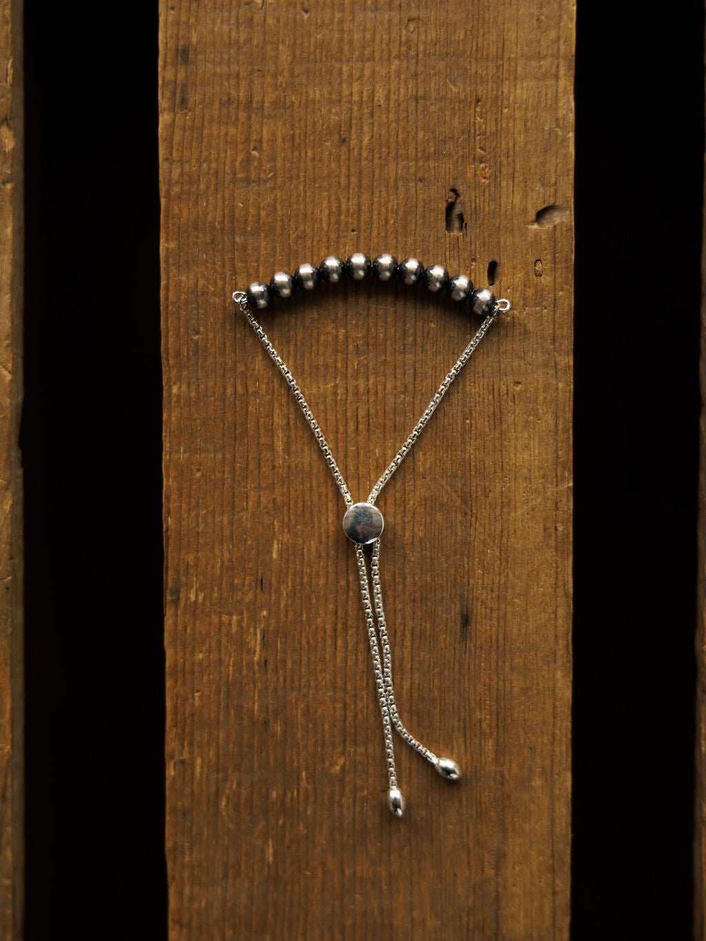 The Elida Bracelet by Savannah Sevens western life{&}style, featuring round dark Navajo pearls and an adjustable sterling silver chain with sliding clasp, is displayed vertically on a rustic wooden surface.