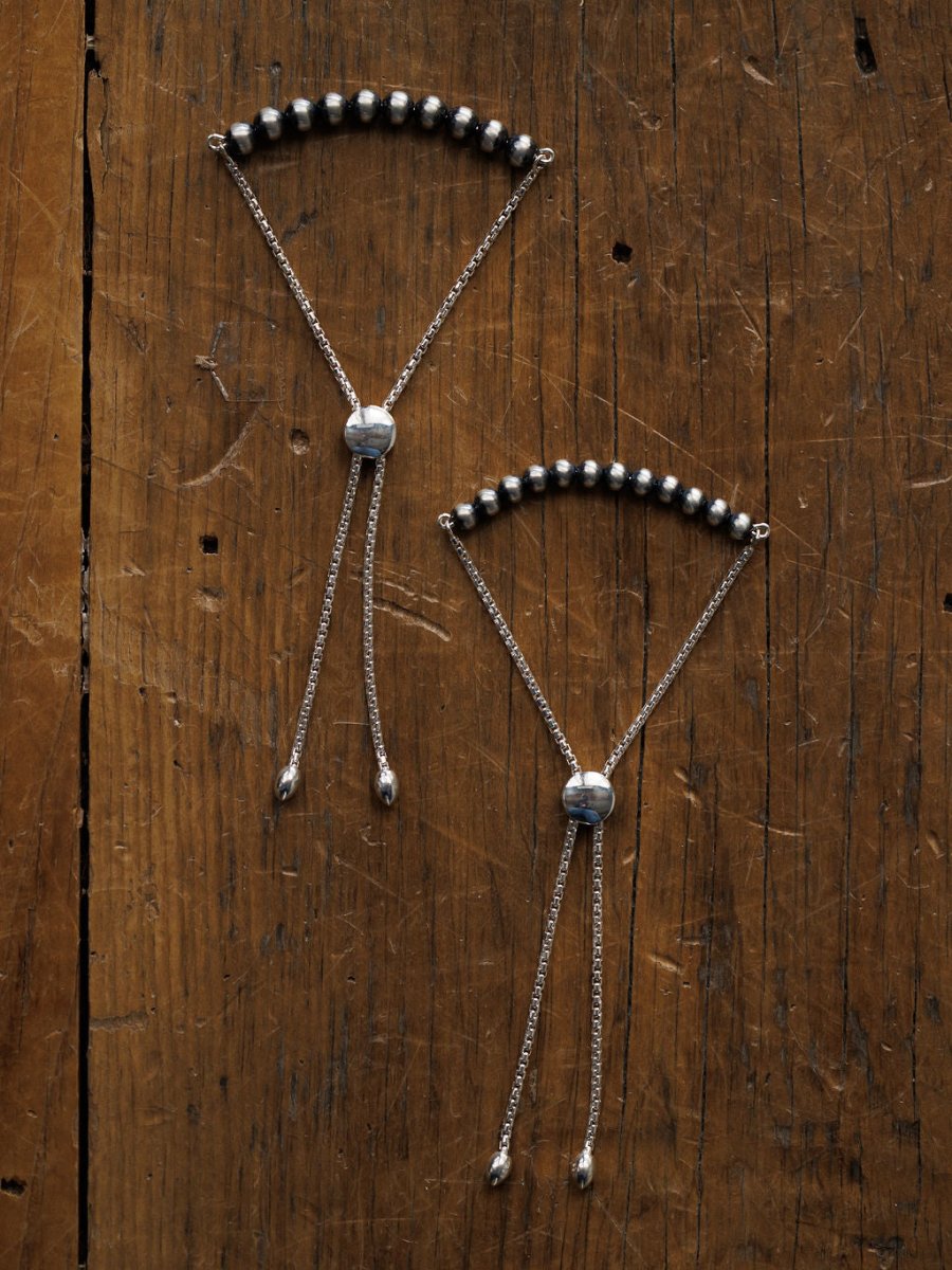 Two Elida Bracelets by Savannah Sevens western life{&}style rest on a rustic wood surface, each with round sterling silver beads and sliding adjustable clasps inspired by classic Navajo pearl designs.