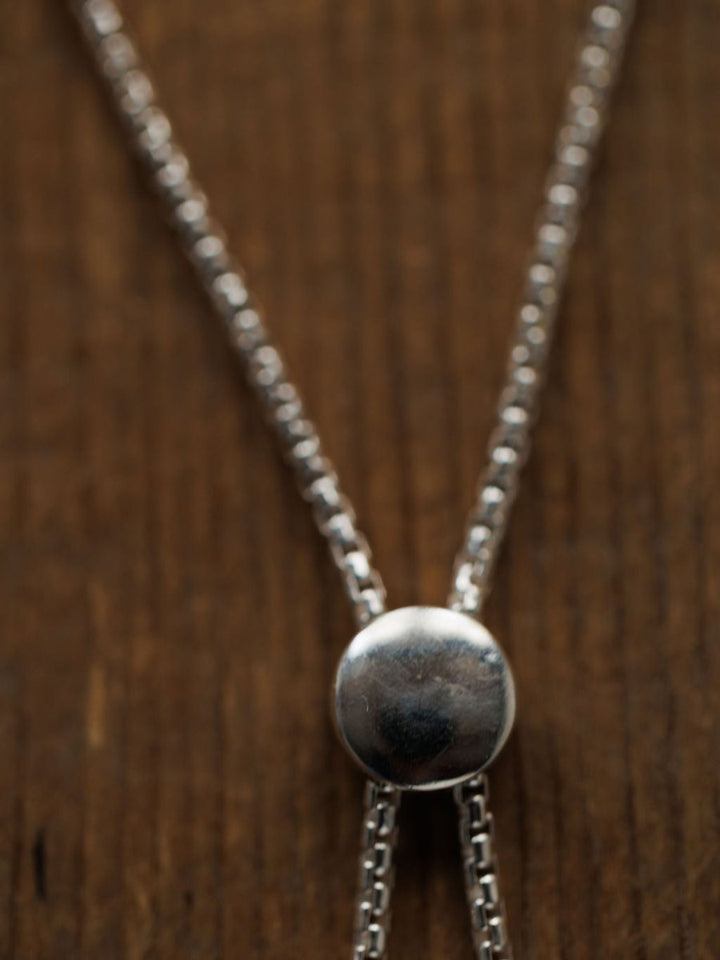 A close-up of a silver necklace with a round pendant and double chains rests on wood beside the Elida Bracelet by Savannah Sevens western life{&}style.