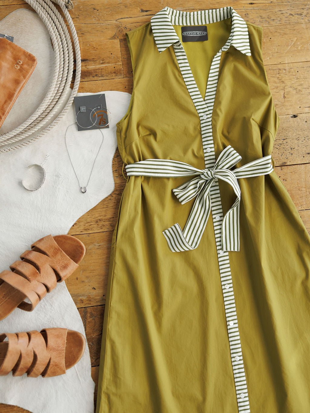 The Elida Chartreuse Striped Dress by Savannah Sevens western life{&}style is displayed on a wooden floor with tan sandals, a silver necklace, a leather bag, rope, and cowhide surrounding it.