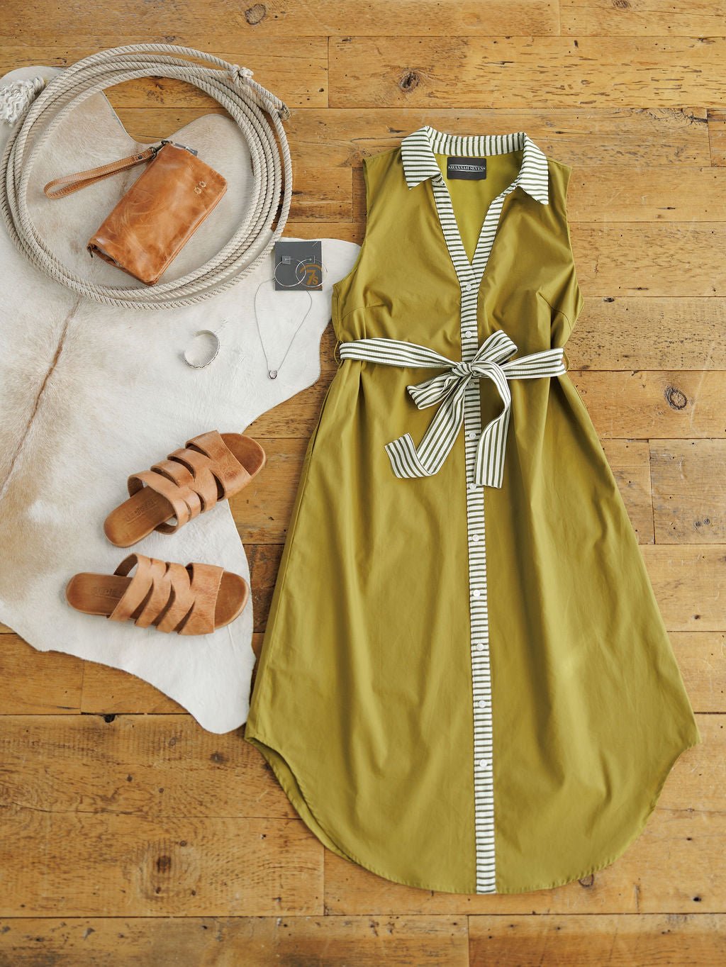 The Elida Chartreuse Striped Dress by Savannah Sevens western life{&}style, a sleeveless midi with white-and-black striped trim and adjustable waist tie, is styled with tan sandals, a brown clutch, silver jewelry, and a rope-accented cowhide rug.
