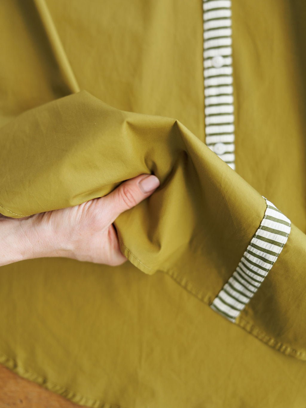 A hand gently holds the Elida Chartreuse Striped Dress by Savannah Sevens western life{&}style, featuring olive chartreuse fabric, white and green striped trim, and an adjustable waist tie.