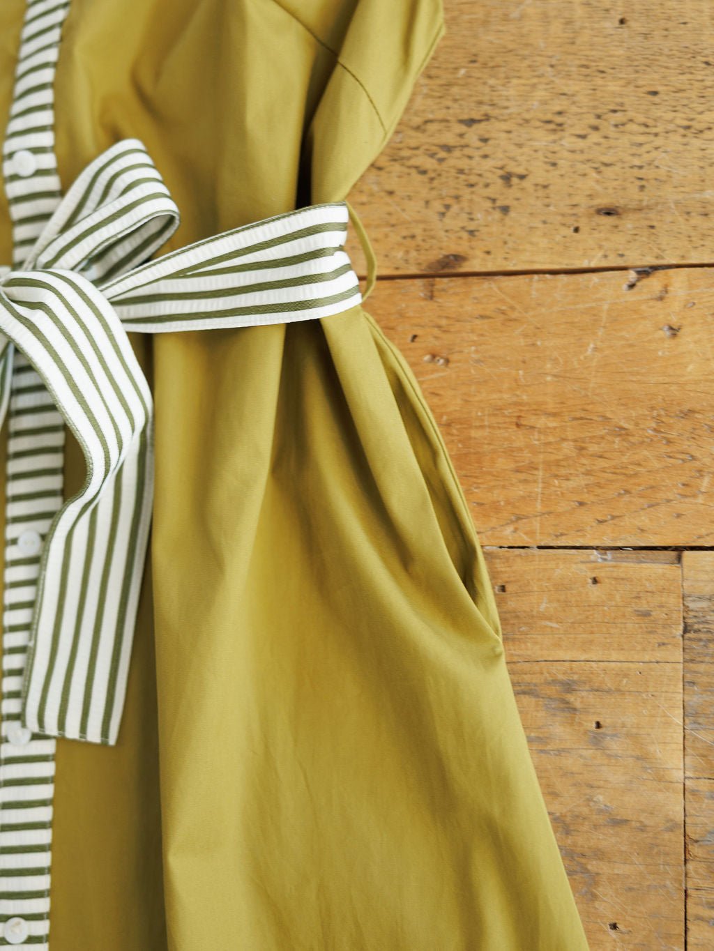 The Elida Chartreuse Striped Dress by Savannah Sevens western life{&}style features white buttons and a wide white-and-green striped adjustable waist tie, shown close-up on a rustic wooden surface.