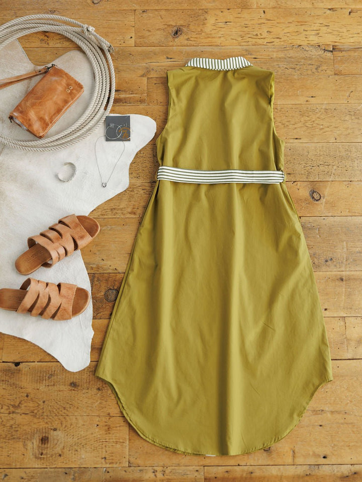 The Elida Chartreuse Striped Dress by Savannah Sevens western life{&}style, a sleeveless midi with striped collar and adjustable waist tie, is styled on a wooden floor with tan sandals, leather purse, jewelry, and mirror atop a cowhide rug.