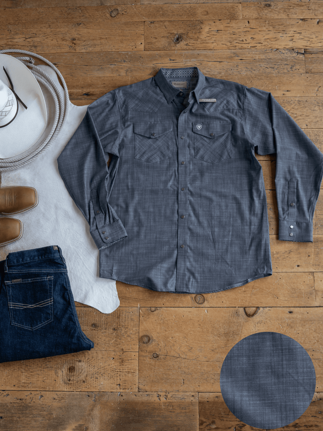 The Elkport Ariat Men's Chambray Button Up by Savannah Sevens western life{&}style in gray is laid flat on a wooden floor beside a white cowboy hat, brown boots, dark blue jeans, and a white cowhide rug. An inset shows its fabric texture.