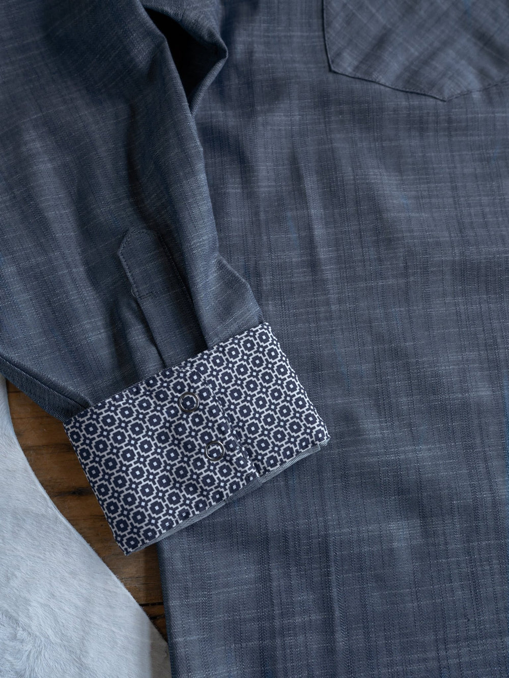 A close-up of the Elkport Ariat Men's Chambray Button Up by Savannah Sevens western life{&}style in blue-gray, featuring a geometric dark blue and white patterned cuff, laid flat on a wooden surface.