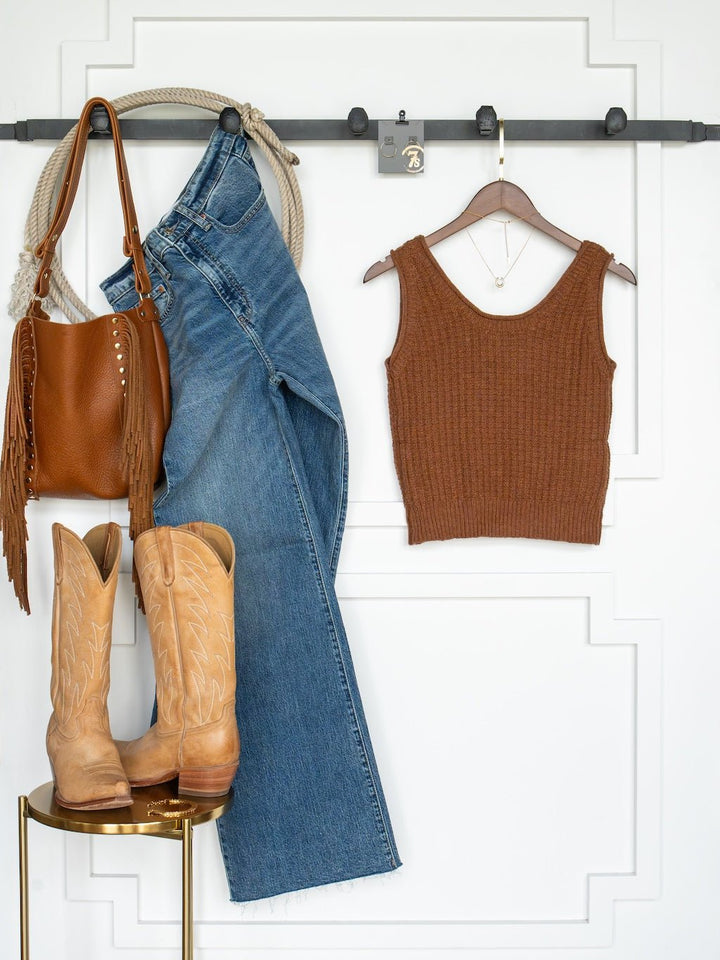 The Ellaville Sienna Knit Sleeveless Top by Savannah Sevens western life{&}style, a pair of jeans, cowboy boots, a brown purse, and a necklace are displayed against a white wall. The clothes hang from hooks while the boots rest on a small round table.