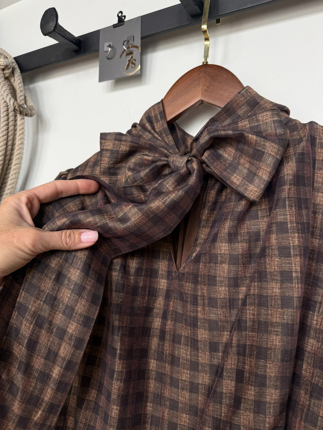 A hand holds the Ellendale Chocolate Gingham Bubble Hem Top by Savannah Sevens western life{&}style, featuring a large bow at the collar, displayed on a wooden hanger against a black wall-mounted rack.