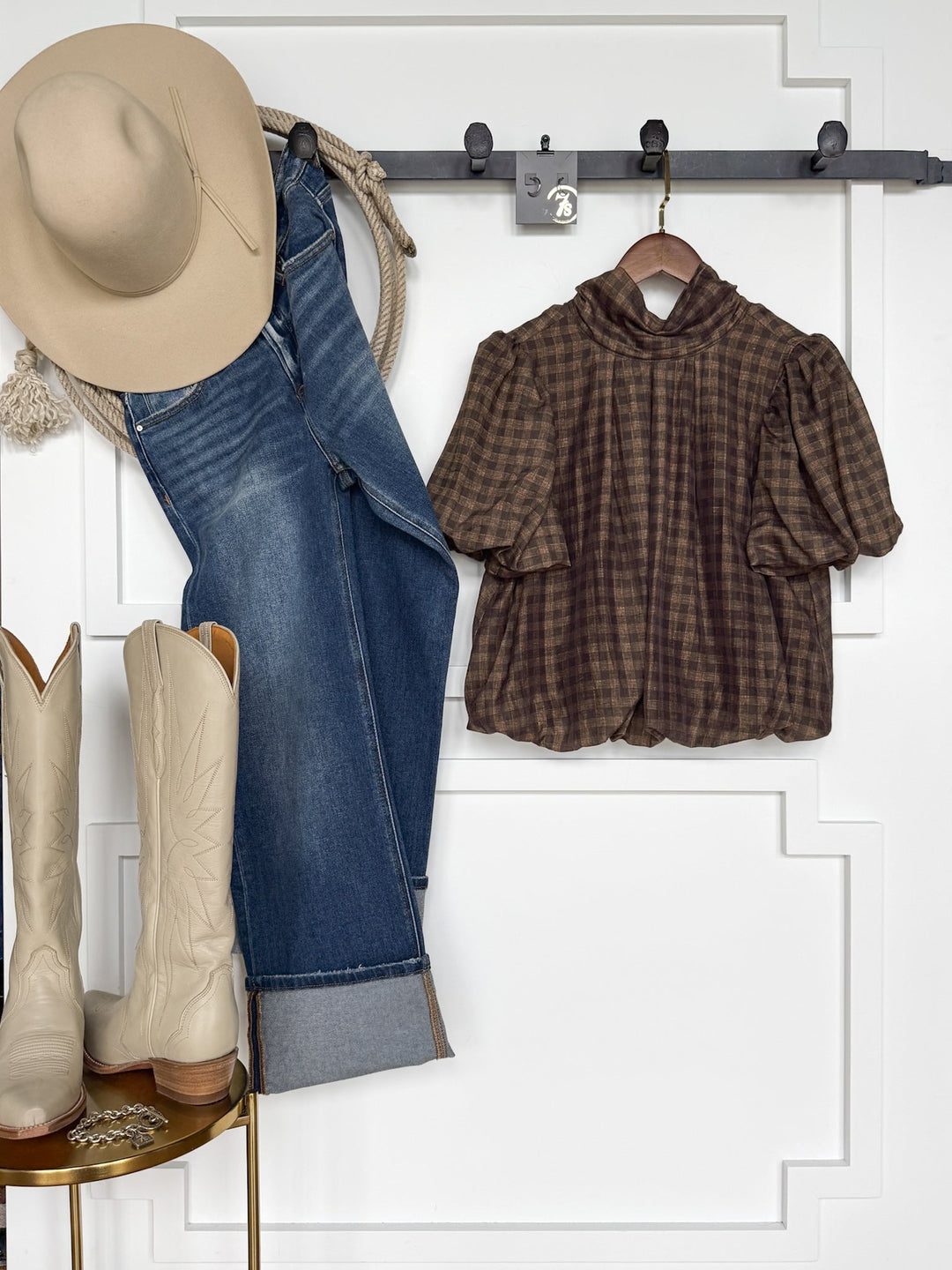 Cowboy hat, rope, blue jeans, the Savannah Sevens western life{&}style Ellendale Chocolate Gingham Bubble Hem Top with short sleeves on hangers, and cream cowboy boots are arranged against a white wall with decorative panel molding.