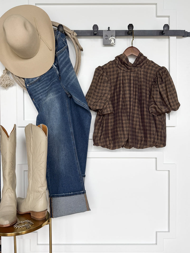 Cowboy hat, rope, blue jeans, the Savannah Sevens western life{&}style Ellendale Chocolate Gingham Bubble Hem Top with short sleeves on hangers, and cream cowboy boots are arranged against a white wall with decorative panel molding.