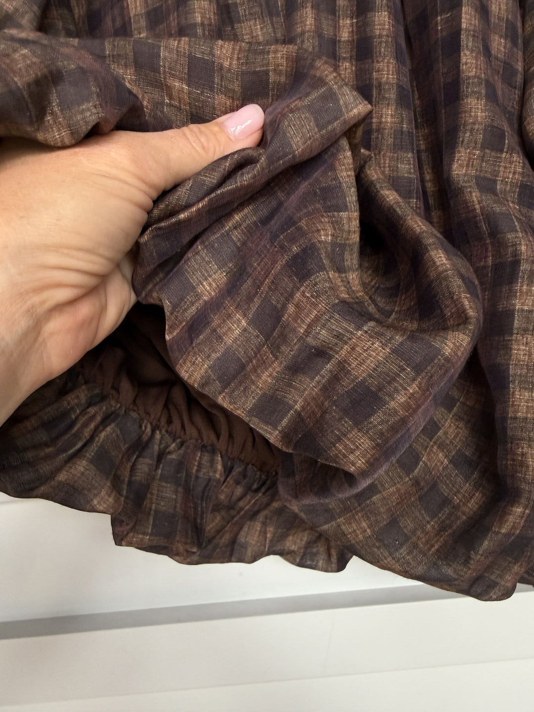 A hand holds the Ellendale Chocolate Gingham Bubble Hem Top from Savannah Sevens western life{&}style, highlighting its brown plaid pattern and gathered elastic seam that creates a signature bubble hem. The texture of the linen blend fabric is clearly visible.