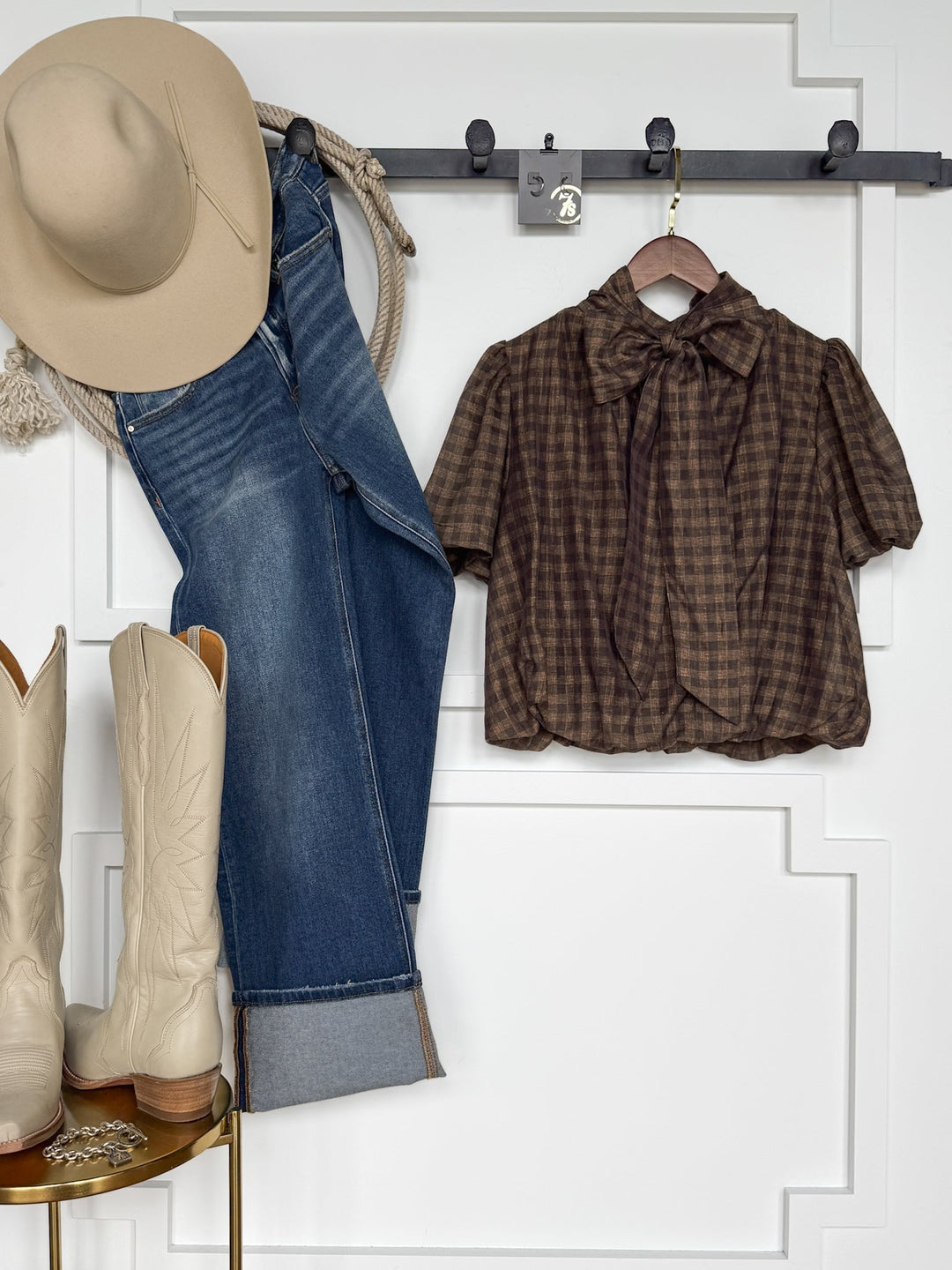 Western outfit: beige cowboy hat, blue jeans, cream cowboy boots, and the Savannah Sevens western life{&}style Ellendale Chocolate Gingham Bubble Hem Top with a bow tie, all displayed against a white wall on hooks and a gold stool.